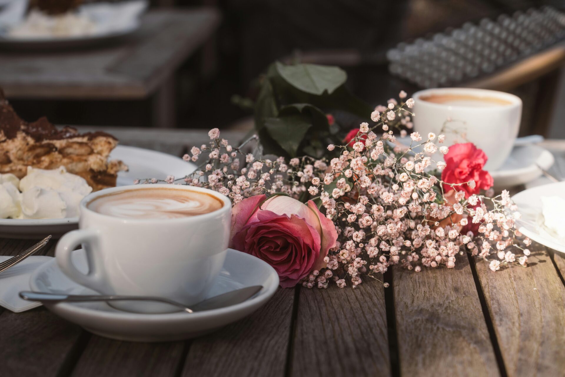 Charmantes Café im Freien mit Cappuccino, Kuchen und Rosen auf einem rustikalen Holztisch.